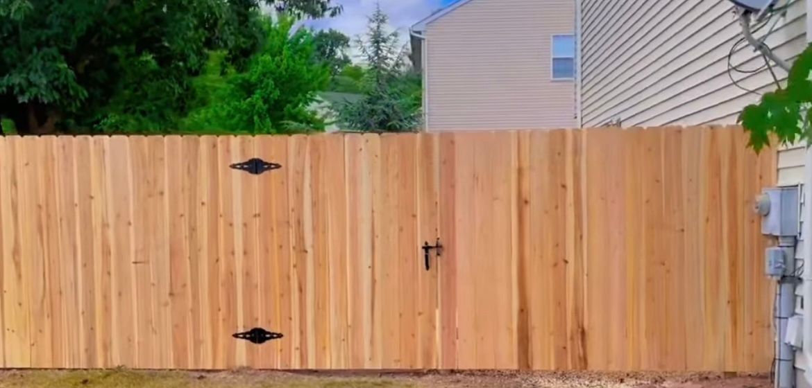 Wooden driveway gate with black hardware in front of a house and greenery