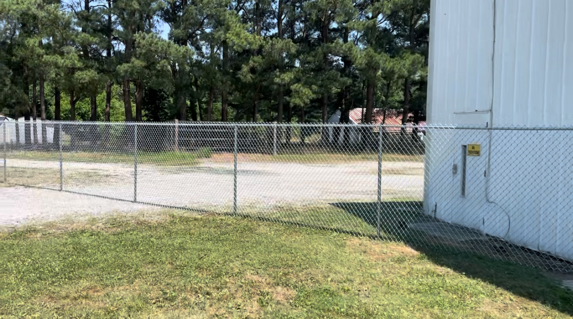 Chain-link fence beside a grassy lot and white building under sunny trees