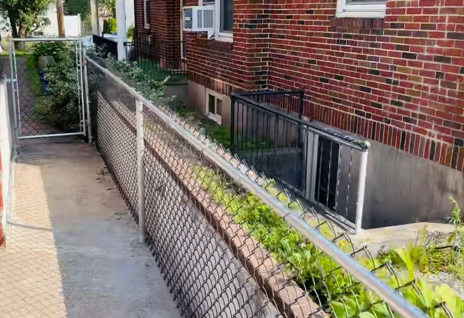 Brick building beside a sloped chain-link fence along a narrow sidewalk with greenery.