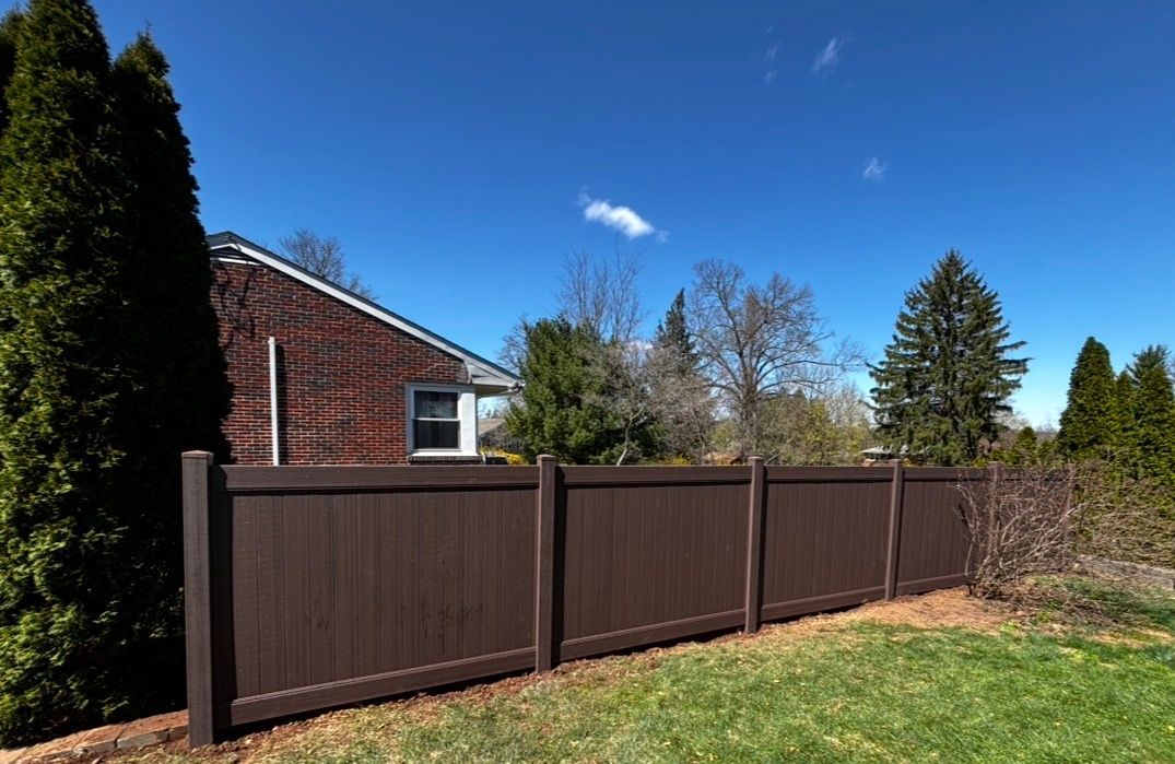 Backyard with brown privacy fence, green lawn, brick house, and trees under a blue sky