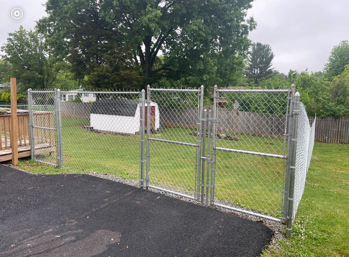 Chain-link fenced grass yard with a white tarp, trees, and a paved path under cloudy skies