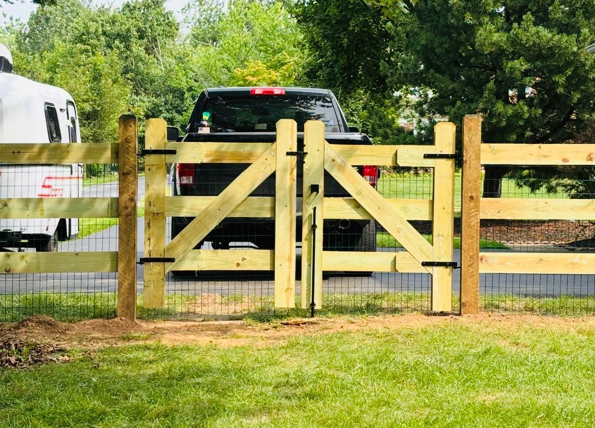 Black pickup truck behind a wooden farm gate in a grassy yard, with trees in the background