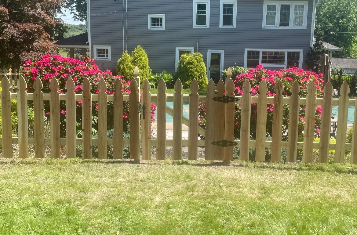 Wooden picket fence in a sunny yard with pink flowers and a gray house in the background