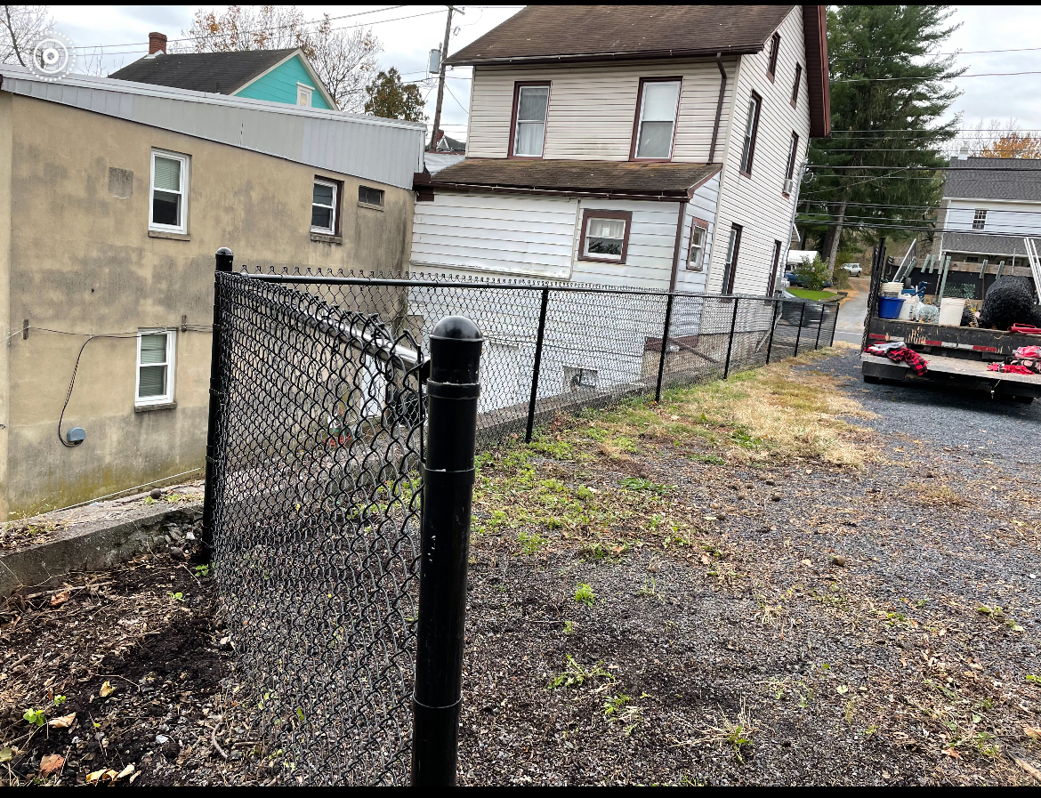 Chain-link fence along a gravel lot beside old houses on a cloudy day, with a parked car in the background