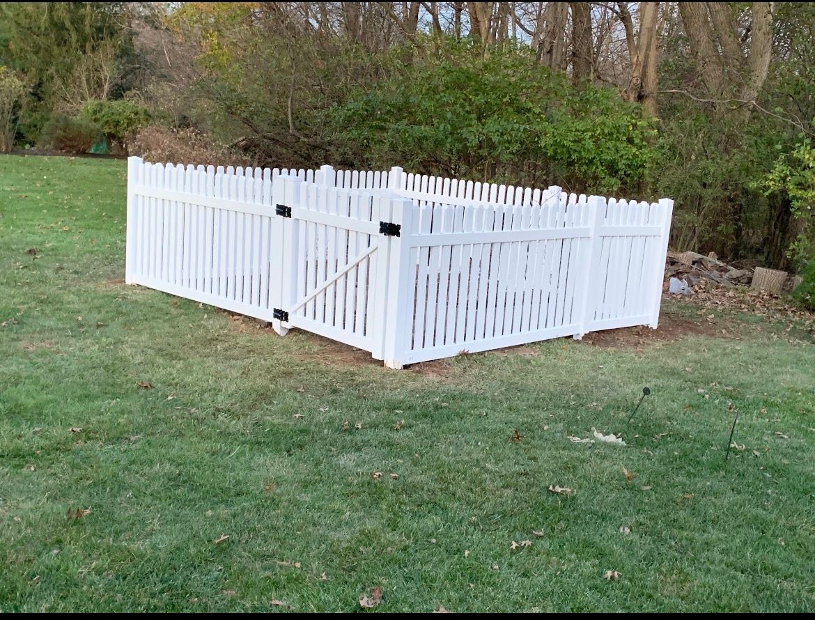 White picket fence enclosure with a gate on a grassy lawn beside trees and bushes