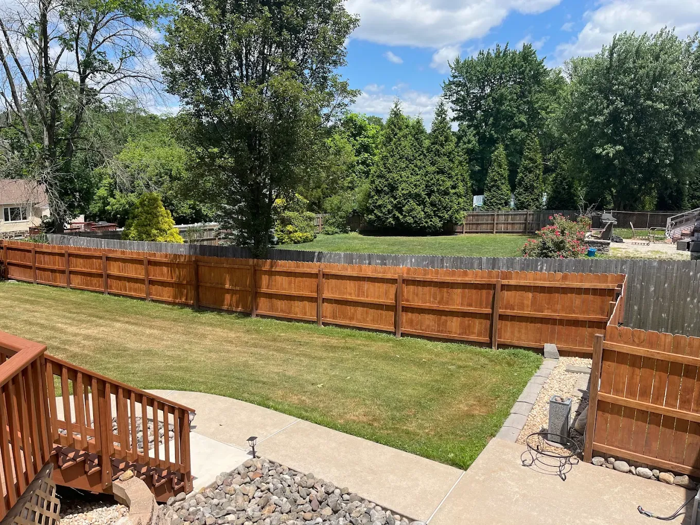 Backyard with wooden fence, green lawn, deck steps, and trees under a partly cloudy sky