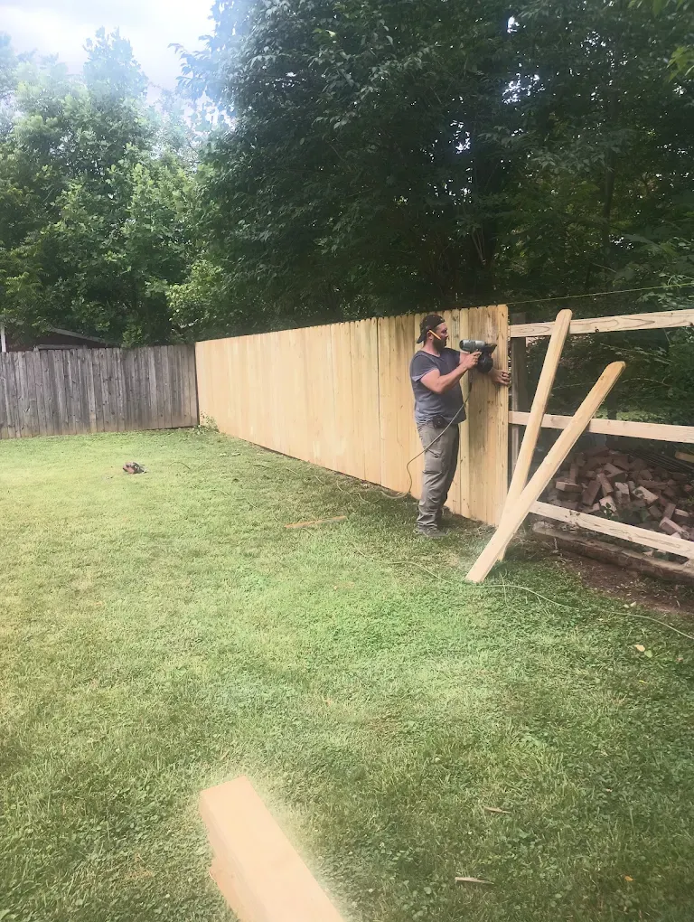 Person building a wooden fence in a grassy backyard, using a red tool against pale boards.