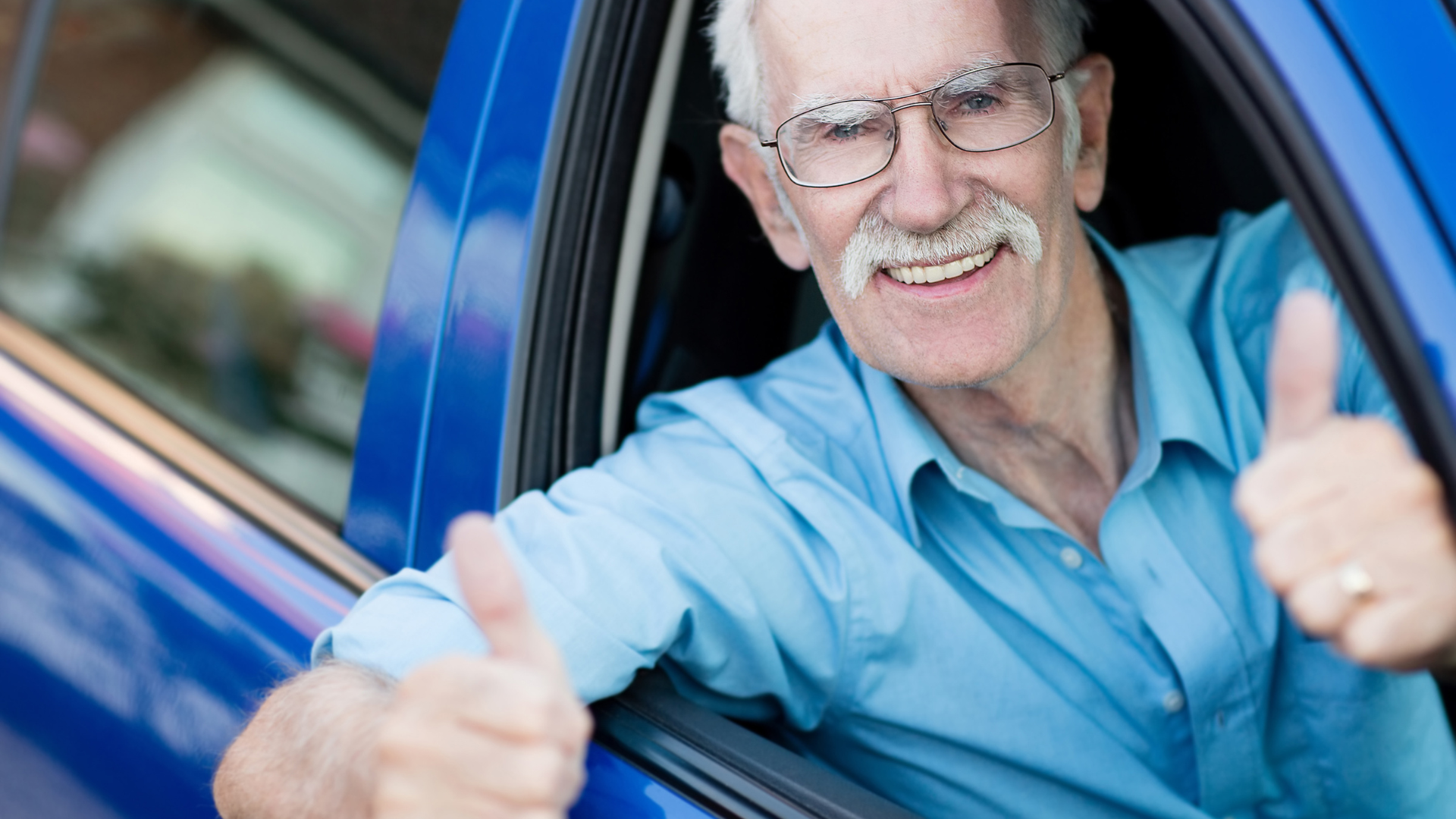 An elderly man is giving a thumbs up while sitting in a blue car.