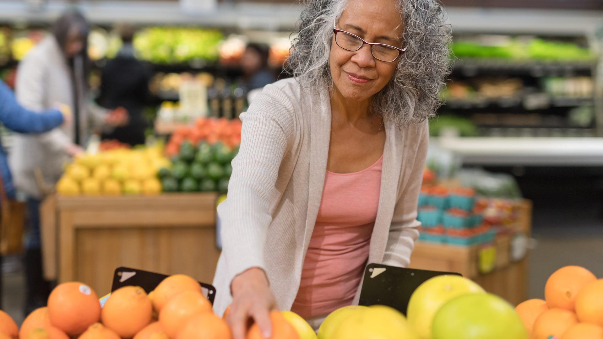 An older woman is standing in front of a fruit stand in a grocery store.