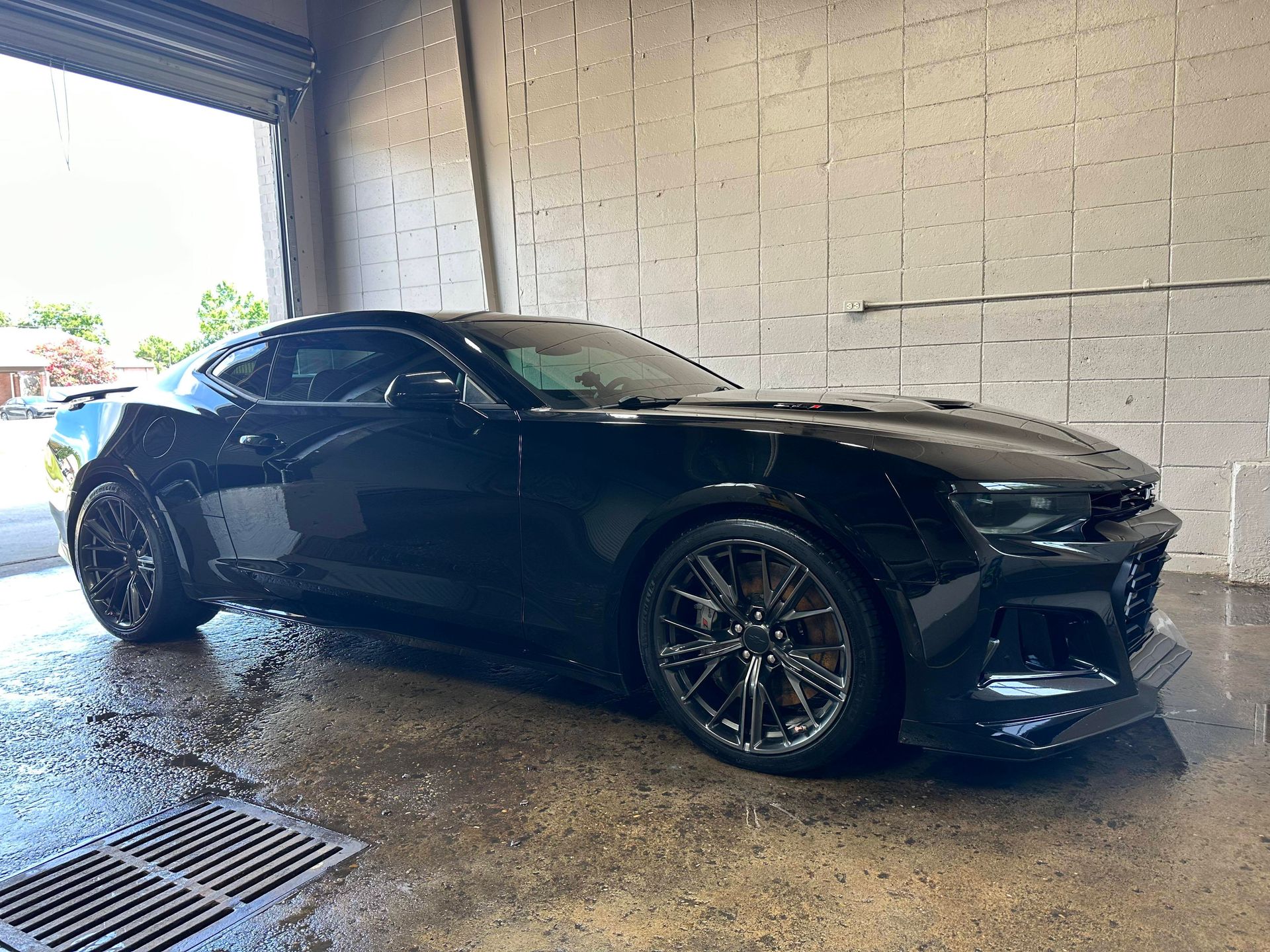 Black sports car being washed inside a garage; wet floor reflects the vehicle and lighting.