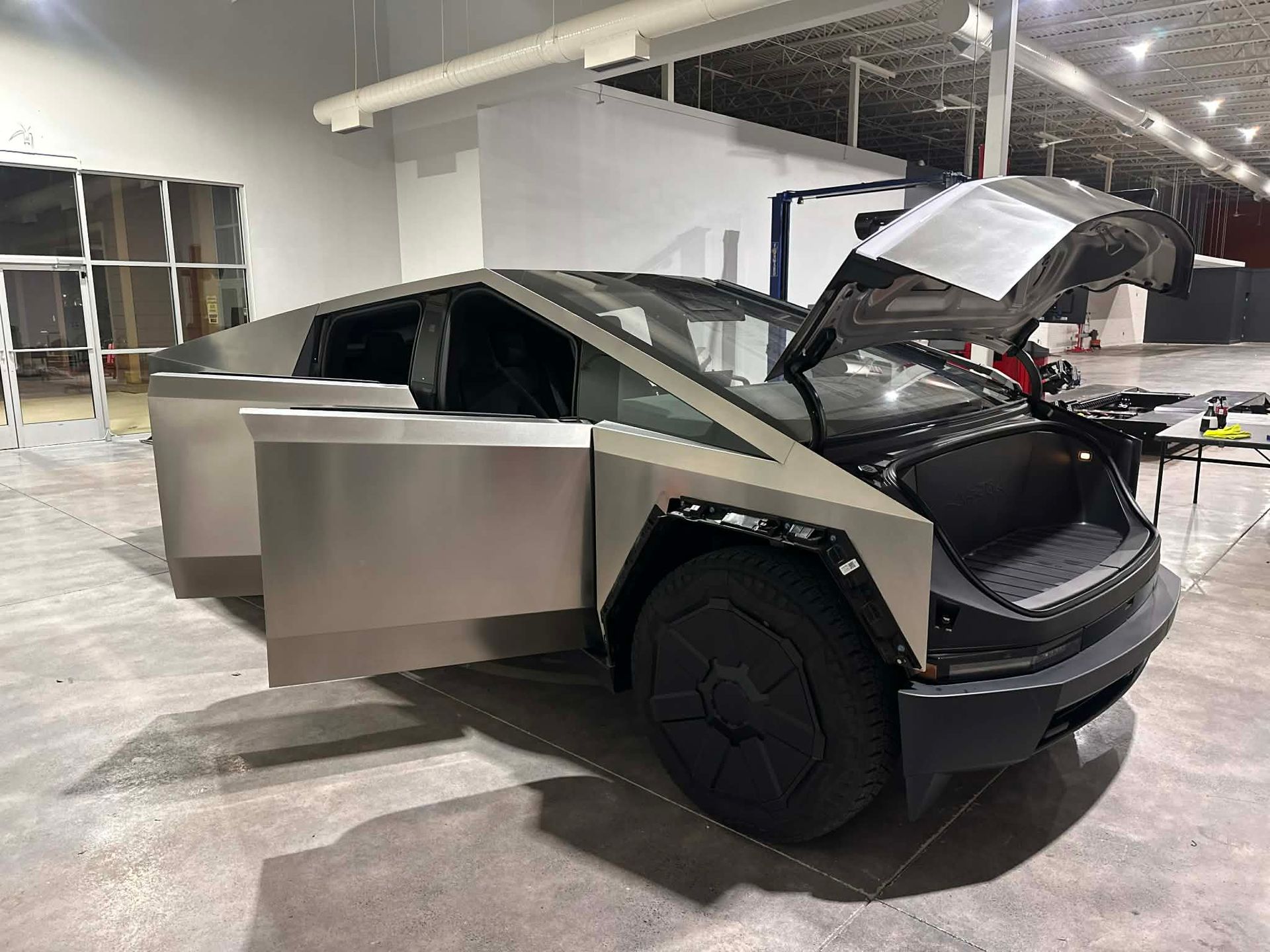 Tesla Cybertruck with doors and front trunk open, indoors. Stainless steel body, black front.