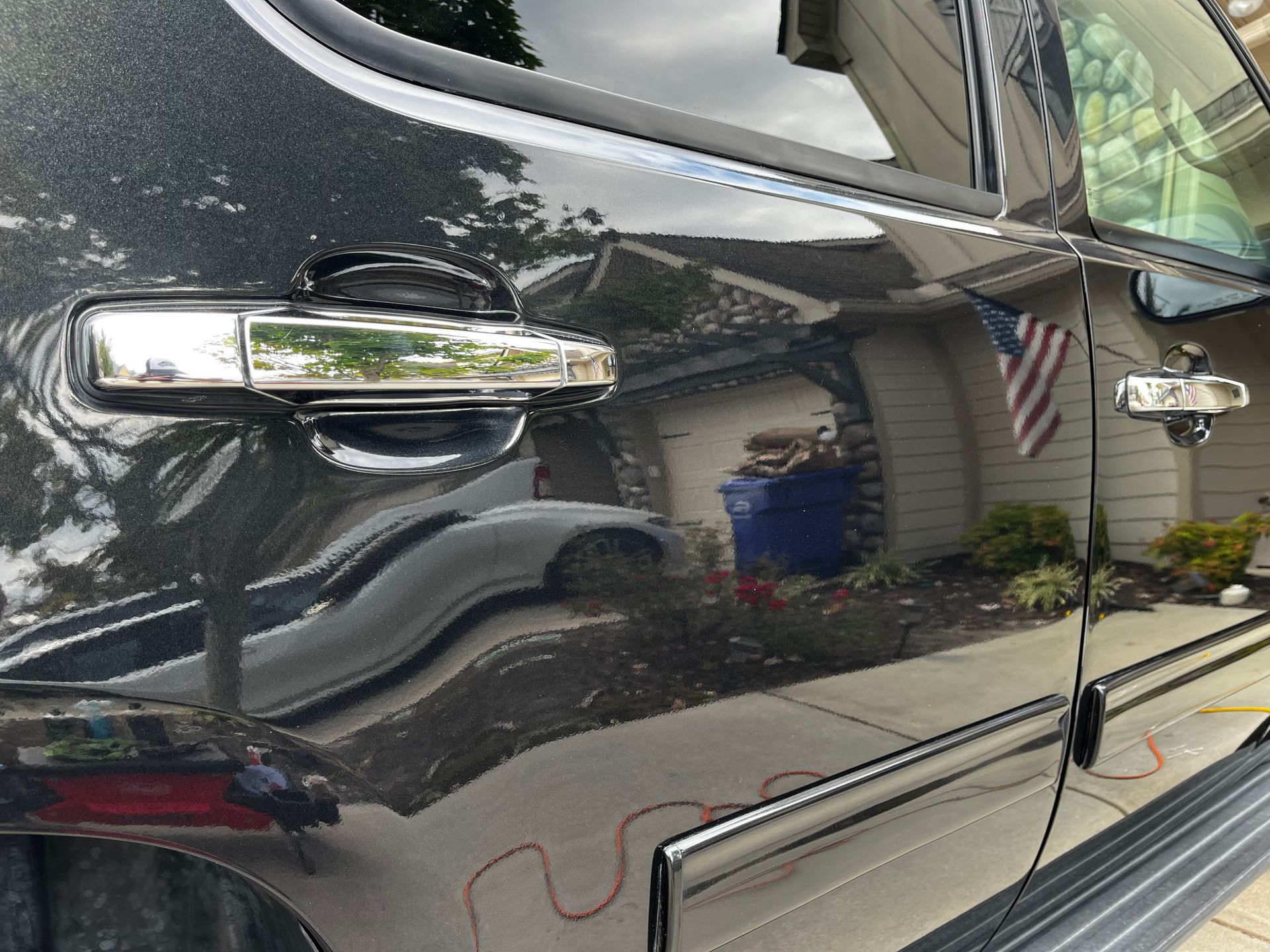 Shiny black truck door with chrome handle, reflecting a house and American flag.