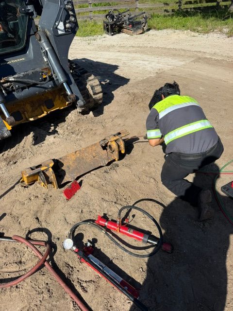 Worker kneeling by hydraulic tools beside excavator on a dirt worksite