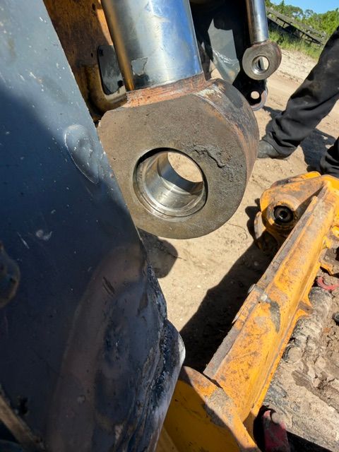 Close-up of a heavy metal excavator joint with a large pin hole and rust, beside a worker’s arm and yellow machinery
