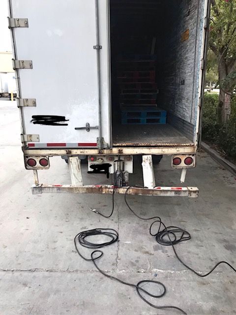 Open rear of a white semi-truck trailer, with pallets visible inside. Cables lie on the ground.