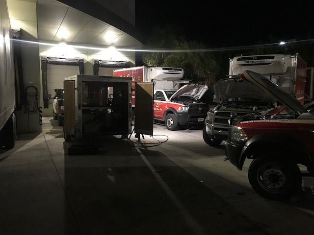 Night scene of several vehicles parked near a building, with hoods open and lights on.