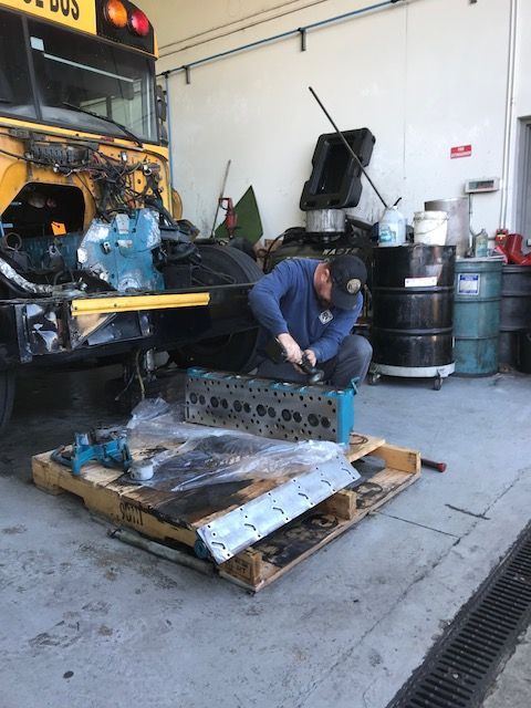 Man working on a bus engine in a garage. He's seated near engine parts on a pallet. A yellow bus is in the background.