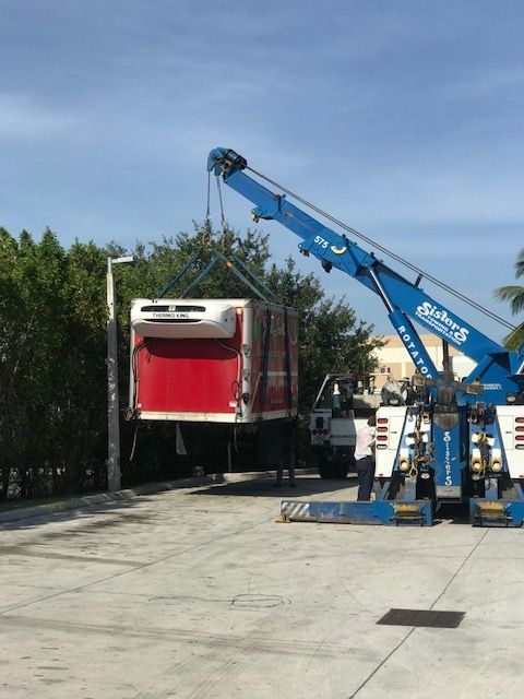 Blue tow truck lifting a red and white refrigerated trailer. Sunny outdoor setting.