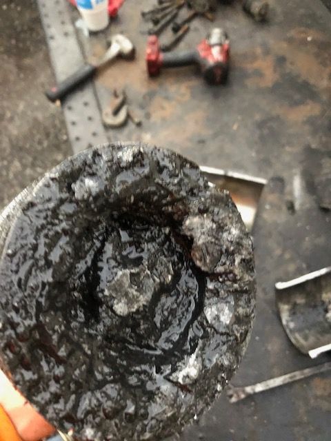 Close-up of a greasy, damaged piston. Black and silver metal with a central depression. Tools are in the blurred background.
