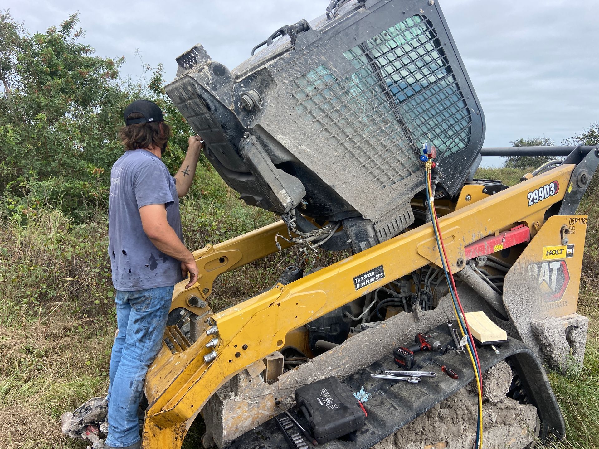 A person working on a yellow skid steer loader with the cab raised in a muddy field.