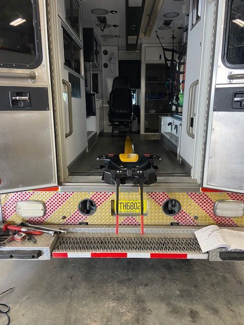 Interior view of an ambulance with rear doors open. A black patient transport system is in place.