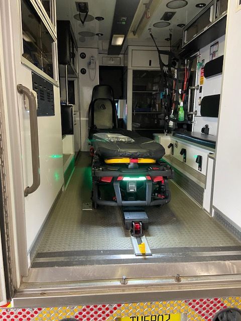 Interior of an ambulance with a stretcher, medical equipment, and storage compartments. White walls, green lights.