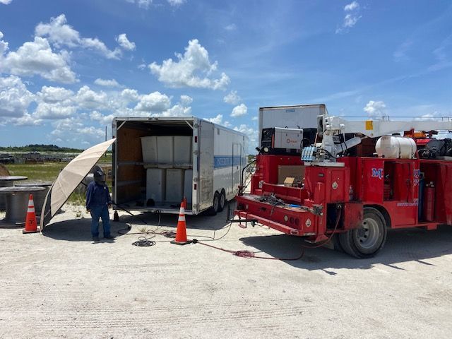 A worker near a white trailer and a red truck with a lift on a gravel lot under a cloudy sky.