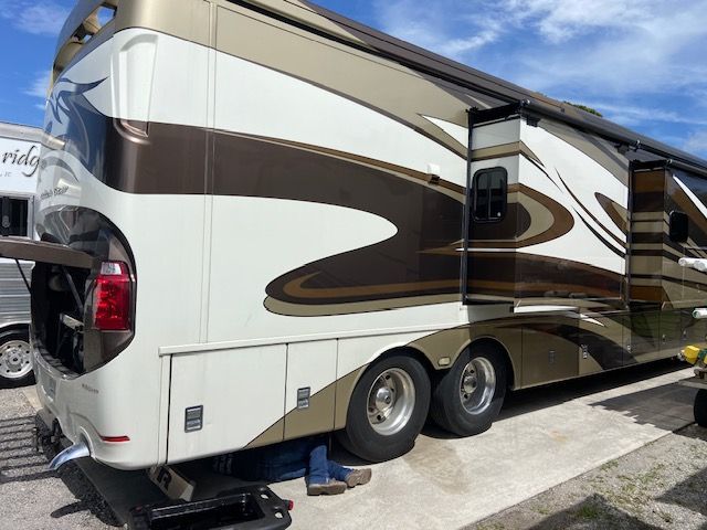 A person works under a large, multi-colored RV. The vehicle is parked outdoors.