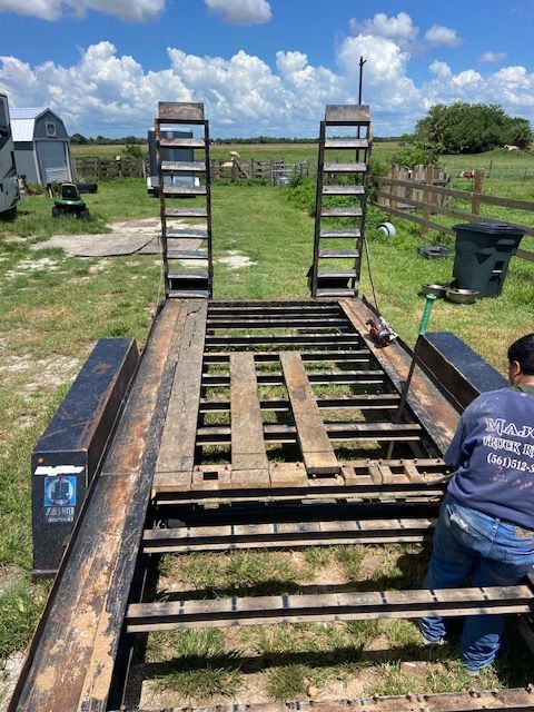Trailer with ramps, partially dismantled, outdoors on grass. Person in blue works on it. Overcast sky.