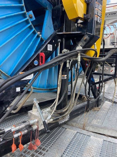 Blue cable reel on a black and yellow machine, with hydraulic hoses and safety tags on a metal platform.