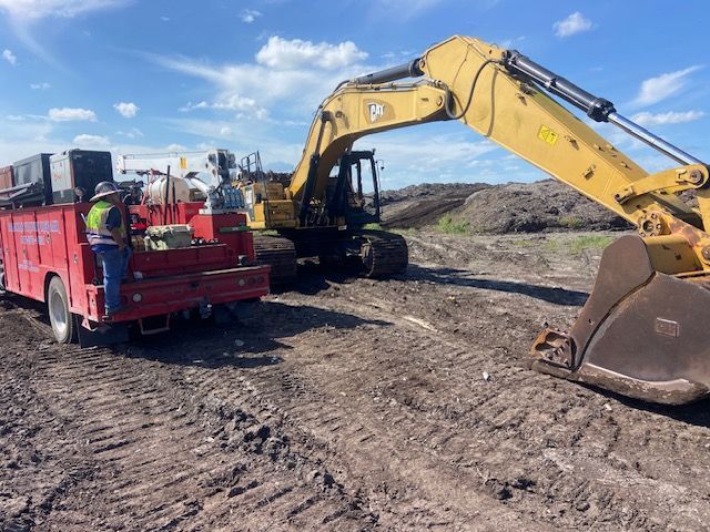 Construction site: yellow excavator, red truck with worker, brown dirt, blue sky.