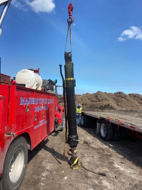 A long, black object suspended by a crane hook, near a red work truck and flatbed trailer.