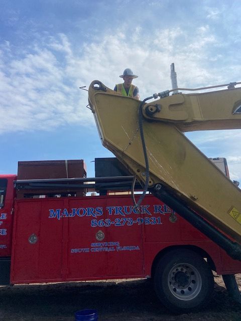 Person in a hard hat on an excavator arm above a red Majors Truck Repair truck under a blue sky.