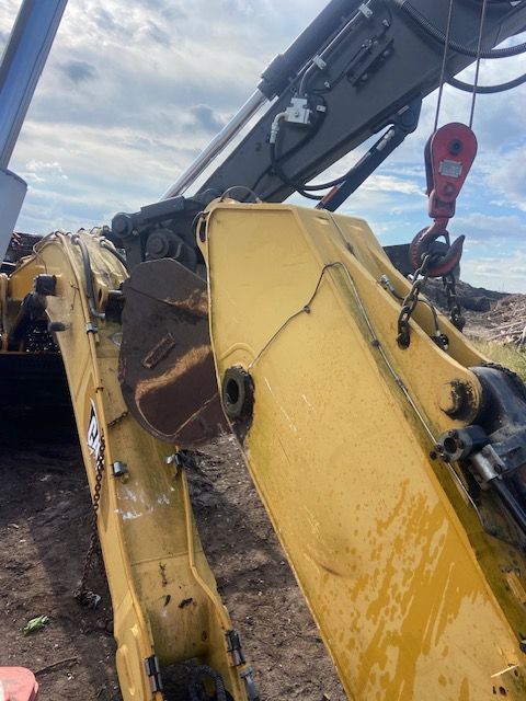 Yellow excavator bucket with dirt, boom, and hook, cloudy sky background.