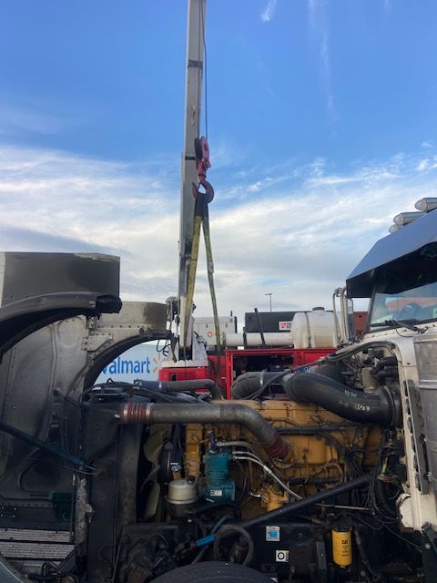 Crane lifting engine from a semi-truck with open hood. Walmart sign in background, cloudy sky.