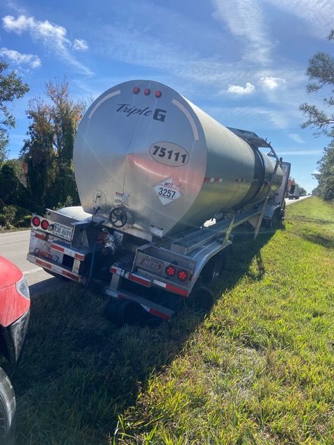 Tanker truck tipped on its side in a grassy ditch beside a road. Silver tank, red truck visible.