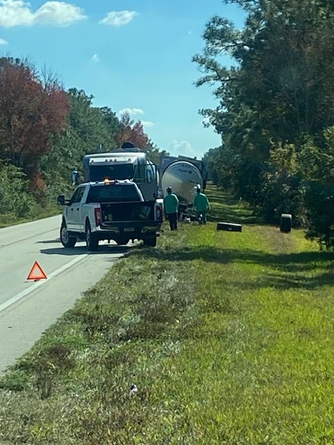 Truck with tank on side of road, workers in green vests, white pickup truck, orange warning triangle.