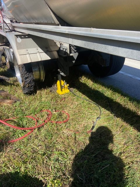 A trailer is being lifted by a yellow hydraulic jack on a grassy area, alongside a road.