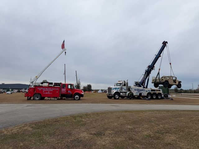 A red crane truck and a white heavy-duty tow truck lifting a tan military-style vehicle in an open, grassy field.