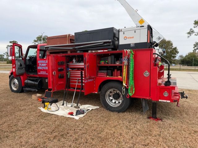 A red utility service truck with open side compartments parked on a field, featuring a crane, tools, and a generator.