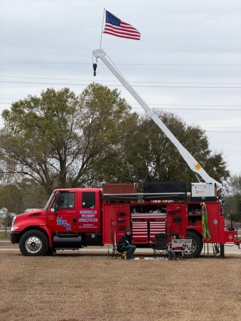 A red service truck with a raised crane and an American flag stands in a field; a person sits nearby under the open door.
