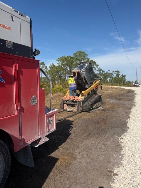 A construction worker in a high-visibility vest operates a yellow skid steer to lift a scrap object on a gravel road.