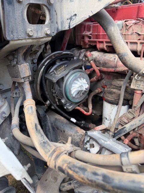Engine bay view of a truck turbocharger with a visible compressor wheel, surrounded by various hoses and engine parts.