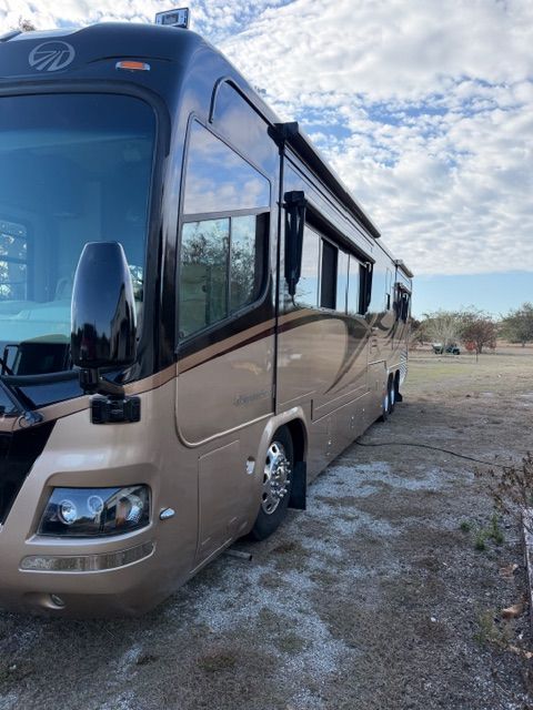 A sleek, bronze and black luxury motorhome parked on a gravel lot under a partially cloudy blue sky.