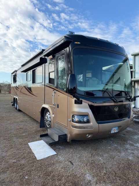 A beige and black motorhome parked on dirt with its entry stairs extended and a small white mat on the ground.