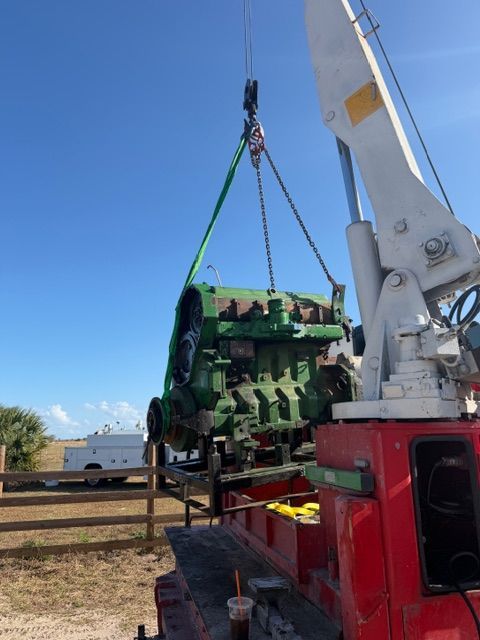 A crane lifts a large, green John Deere engine block outdoors on a sunny day.