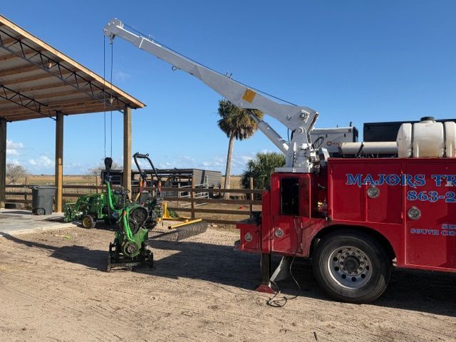 A red utility truck with a crane attachment lifting a green piece of farm equipment outdoors under a wooden pavilion.