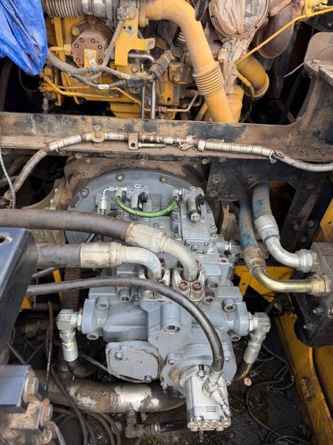 The engine bay of heavy machinery shows a large, grey hydraulic pump connected to multiple hoses beneath a yellow engine.