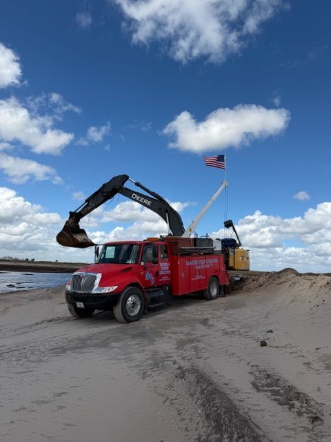 A red utility truck with an extended crane arm parked on a sandy beach under a blue sky with an American flag.