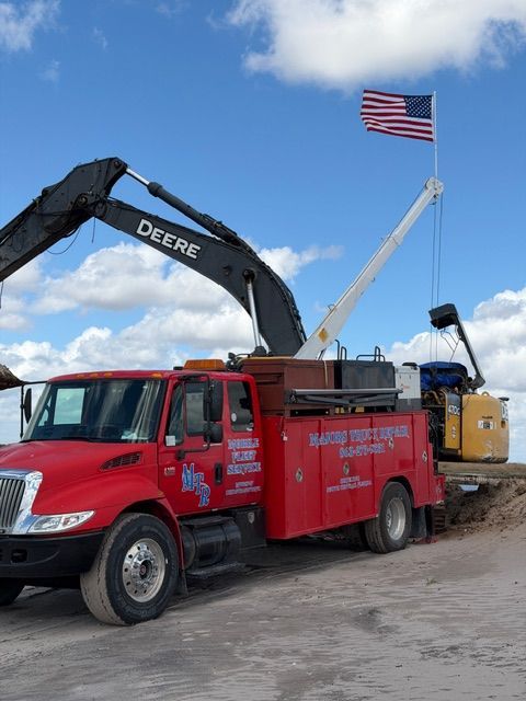 A red service truck with a boom crane and a mounted American flag parked on a dirt lot under a blue, cloudy sky.